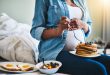 Schwangere Frau mit Pfannkuchen auf dem Schoss, daneben eine Schüssel mit Früchten und ein Teller mit Toast und Spiegelei