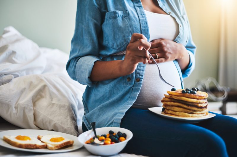 Schwangere Frau mit Pfannkuchen auf dem Schoss, daneben eine Schüssel mit Früchten und ein Teller mit Toast und Spiegelei