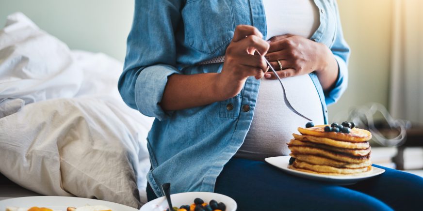 Schwangere Frau mit Pfannkuchen auf dem Schoss, daneben eine Schüssel mit Früchten und ein Teller mit Toast und Spiegelei