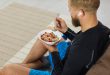 Junger Sportler sitzt auf Sofa und isst Granola