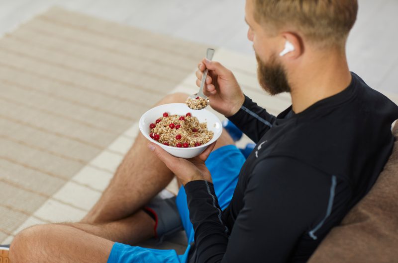 Junger Sportler sitzt auf Sofa und isst Granola