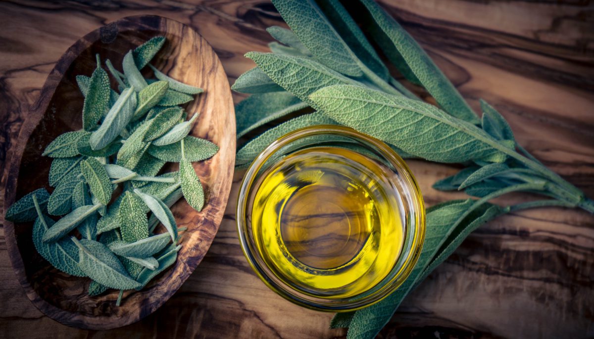 Sauna und Aromatherapie verbinden Wärme und Duft zu einem ganzheitlichen Erlebnis, das Körper und Geist gleichermaßen belebt Salbeiblätter in Holzschale und auf Holztisch neben Glas mit Öl - Saunaaufguss selber machen – Rezepte für tollen Duft aus wunderbaren Ölen